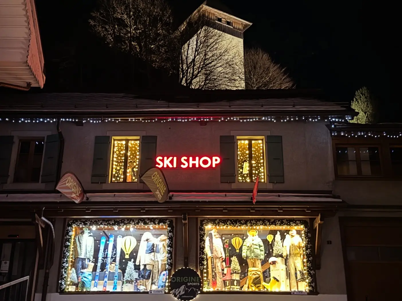 Façade illuminée du magasin Origine Sports à Château-d’Œx la nuit, avec l’enseigne Ski Shop et les vitrines décorées pour l’hiver